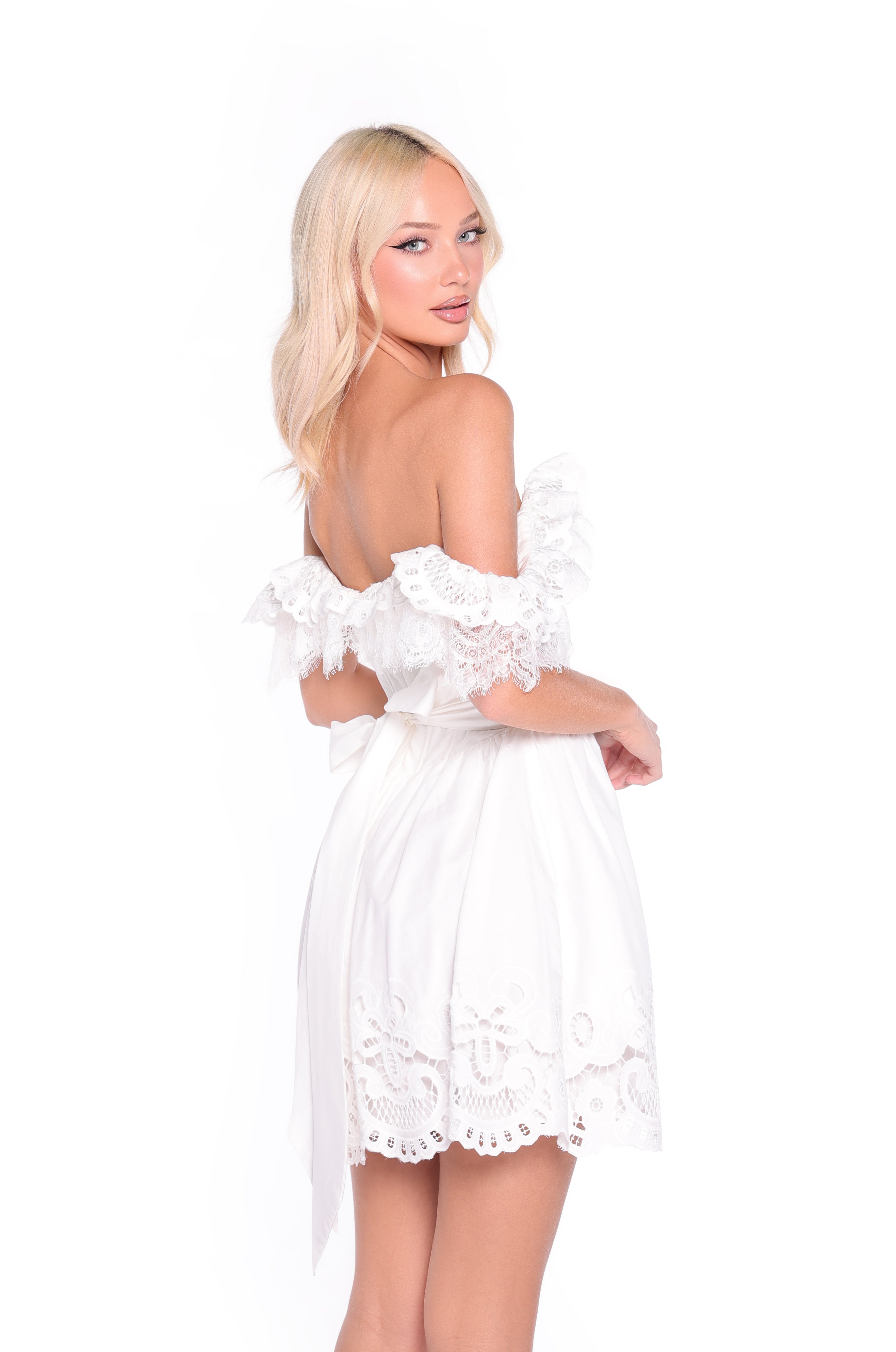 A woman with long blonde hair wears the Ari cap-sleeve short ivory graduation dress and poses against a plain white background, looking over her shoulder.